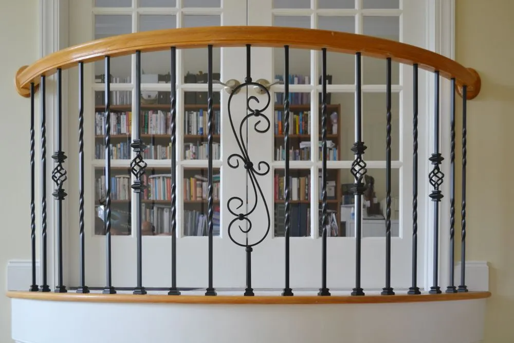 Wooden and wrought-iron balcony railing with curved top rail. Bookshelves visible through the window behind it.