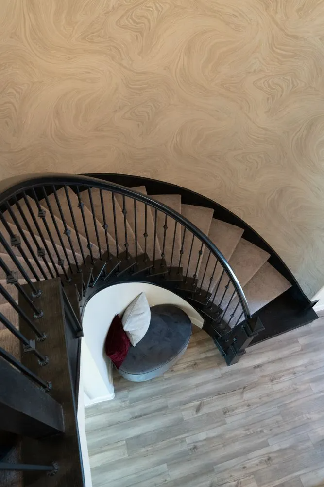 Curved staircase with black railing, beige carpeted steps, and wood-look flooring. A small seating area is below.