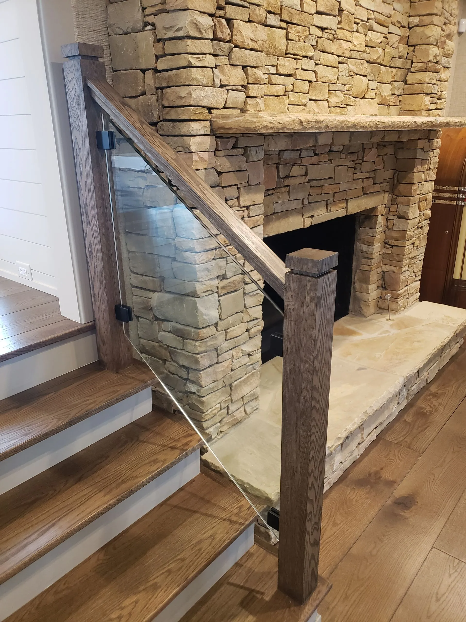 Staircase with glass railing, stone fireplace in the background, hardwood floors.