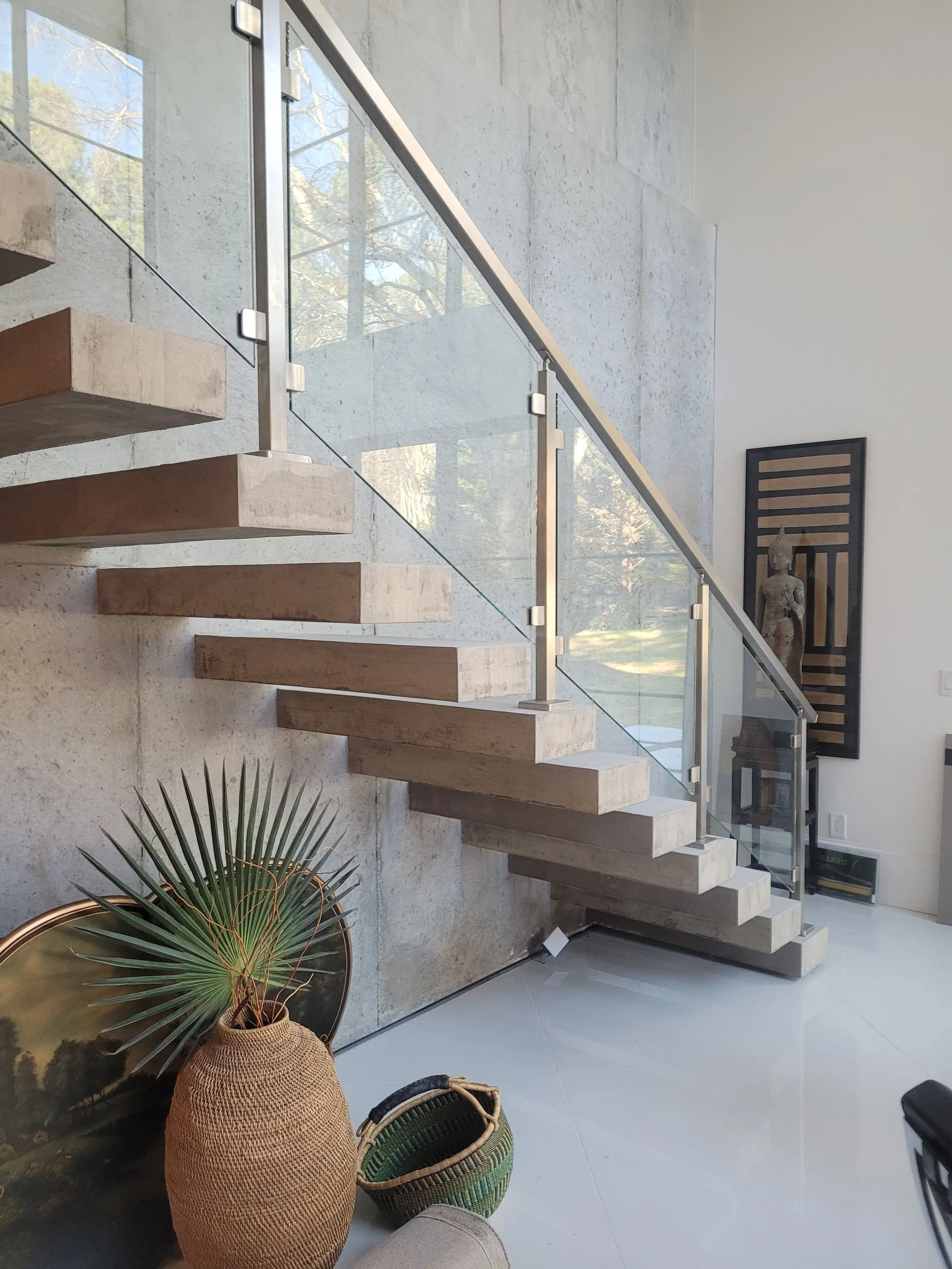 Modern staircase with concrete steps, glass railing, and textured wall. Decorative plants and art in the foreground.