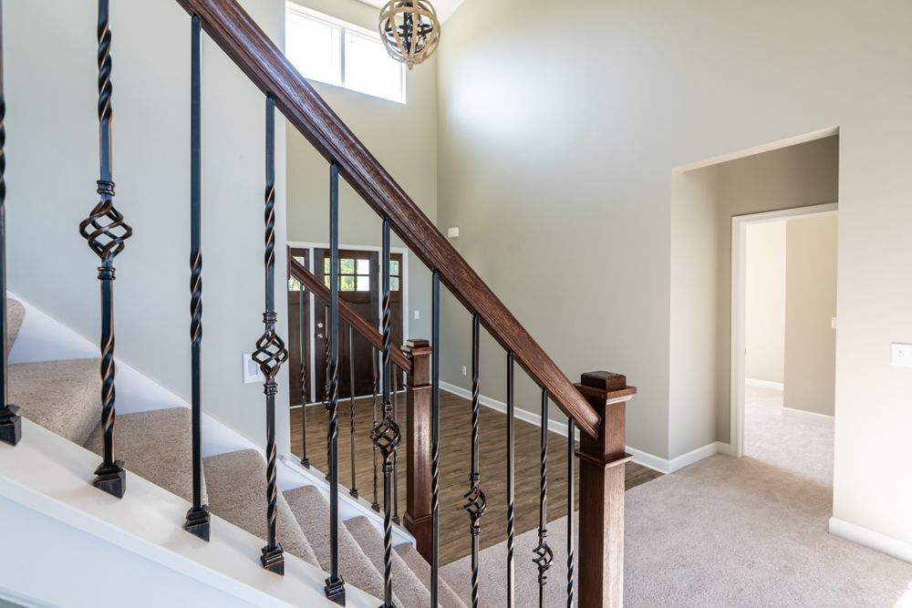 Staircase with brown wooden railing, black iron balusters, leading up to a foyer with a front door and carpet flooring.