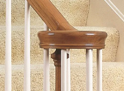 Close-up of a wooden stair railing, focusing on the newel post and balusters. Light brown wood against beige carpet.