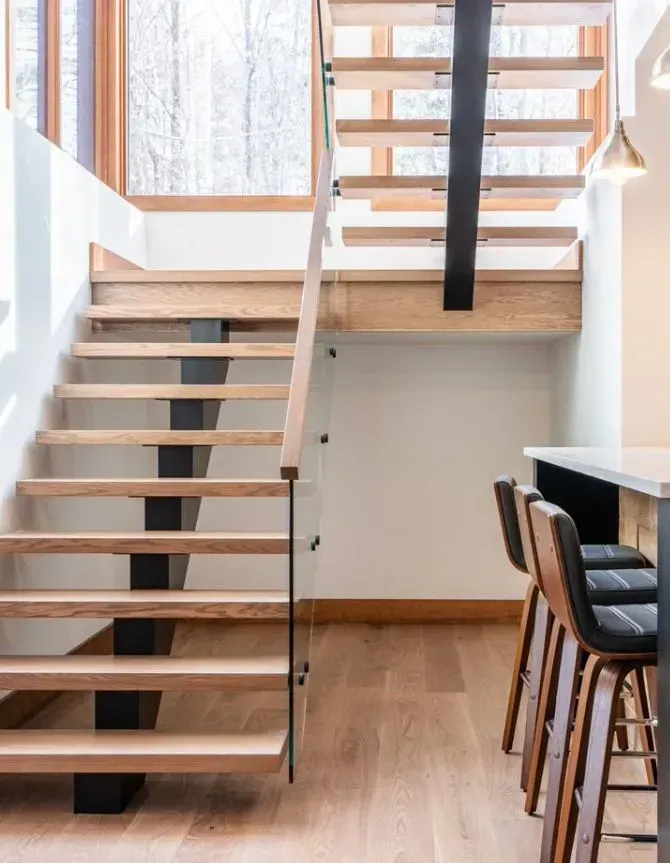 Wooden staircase with black supports and glass railing next to a kitchen with bar stools.
