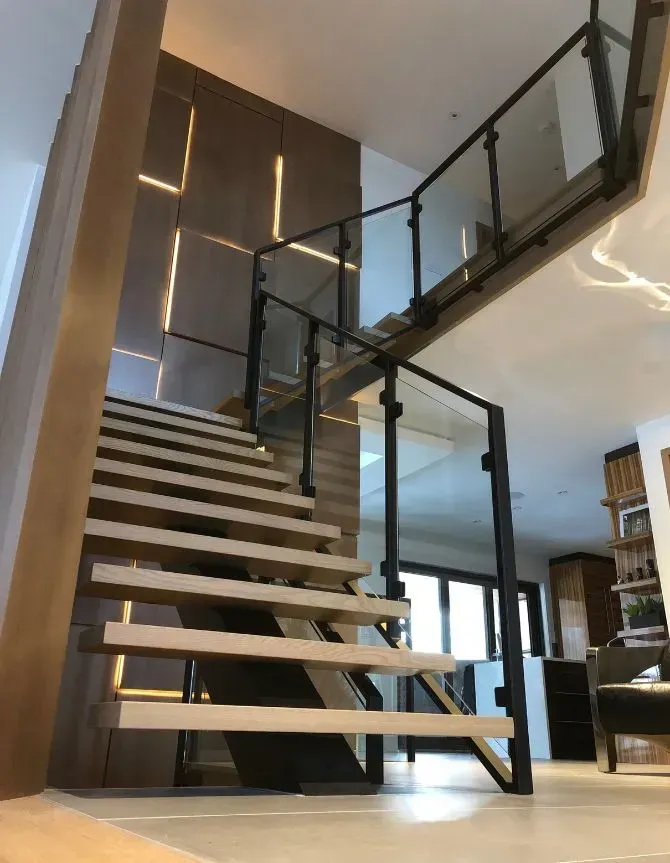 Modern wooden staircase with glass railings and black supports, ascending to a second floor. Interior with accent lighting.