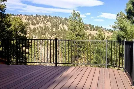 Wooden deck with black railings overlooking a forested hillside under a blue sky.