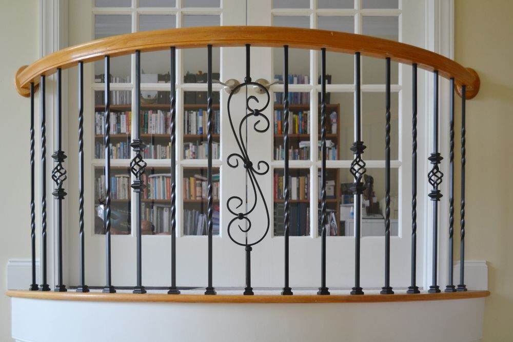 Balcony railing with black metal spindles and wooden handrail, arched, in front of a white door with bookshelves.