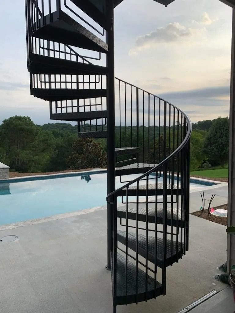 Black spiral staircase next to a pool, overlooking a lush green landscape.