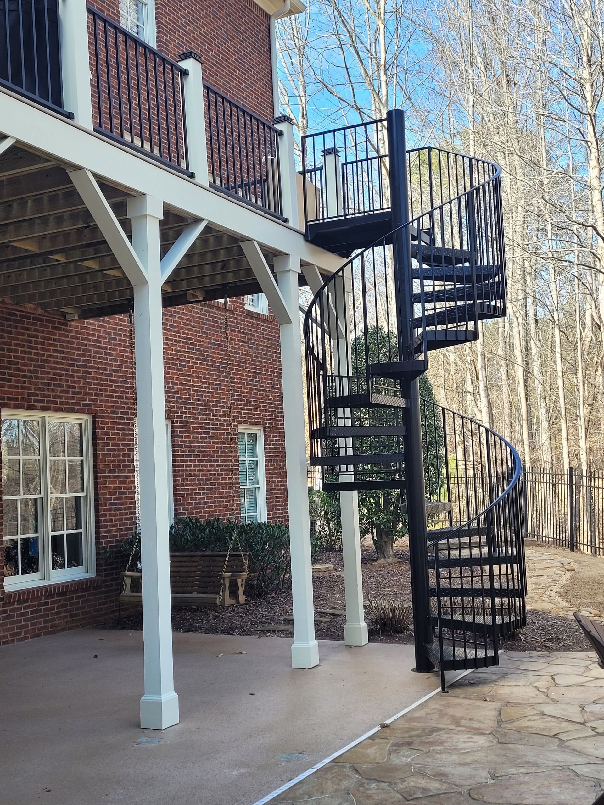 Black spiral staircase leading from a white-columned deck to a concrete patio. Brick building and trees in the background.