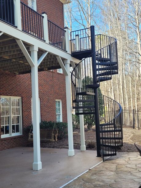 Black spiral staircase leading from a white-columned deck to a concrete patio. Brick building and trees in the background.