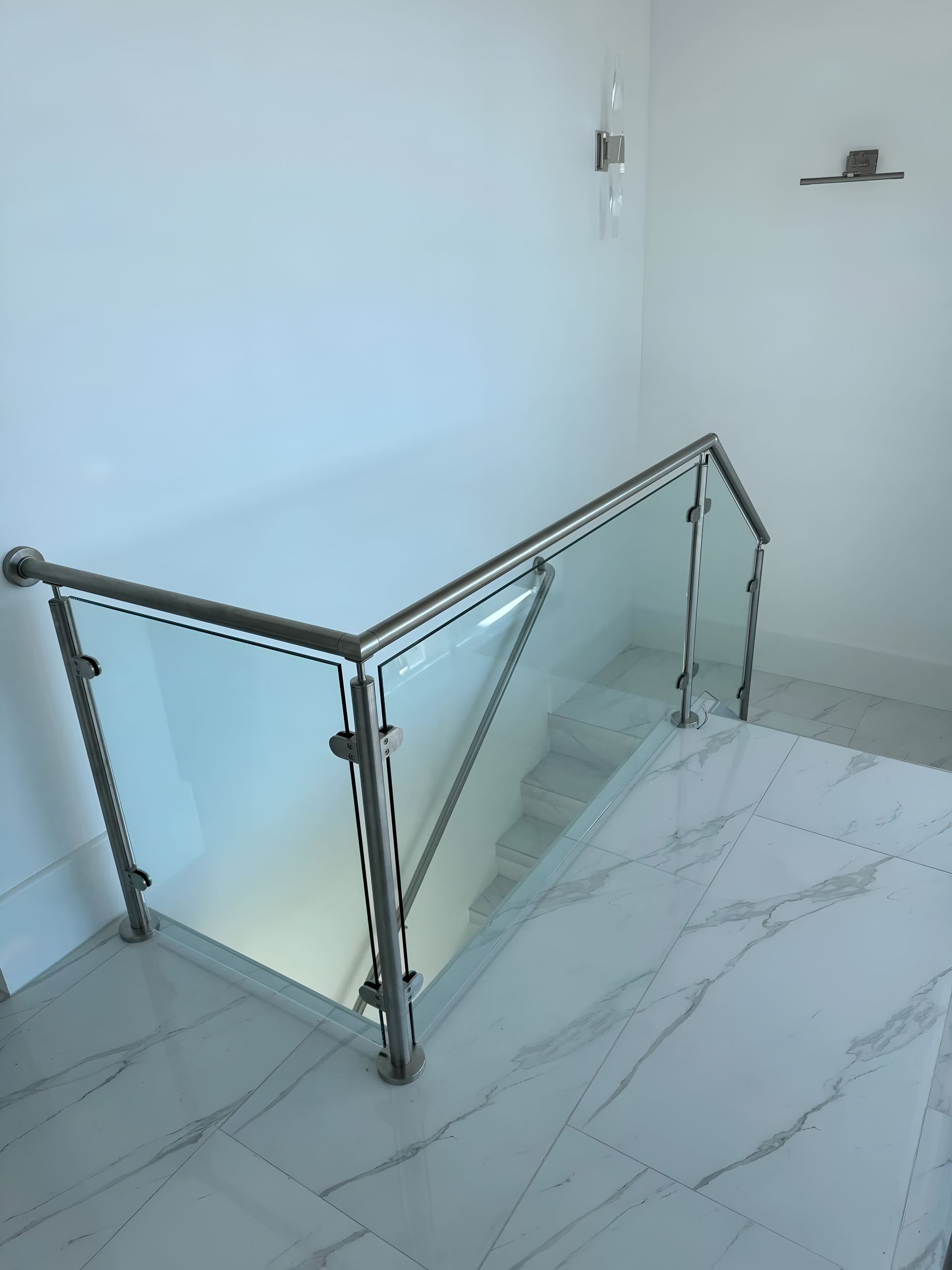 Glass and stainless steel railing on a staircase with white walls and marble flooring.