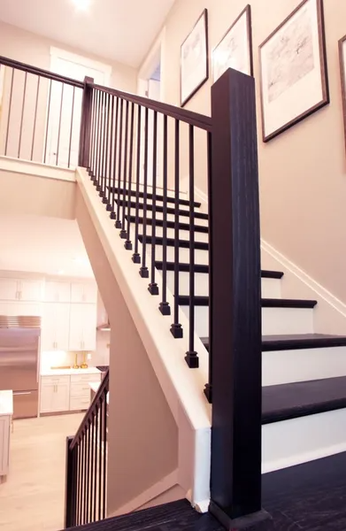 Staircase with black railings and steps, white risers, and a light beige wall. Kitchen visible in the background.