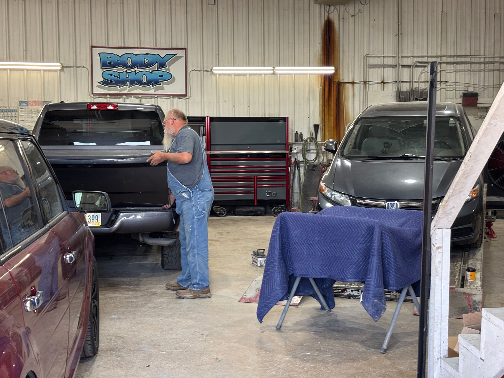 A man in overalls installing a truck bed cover inside a garage. Gray truck and car are present.