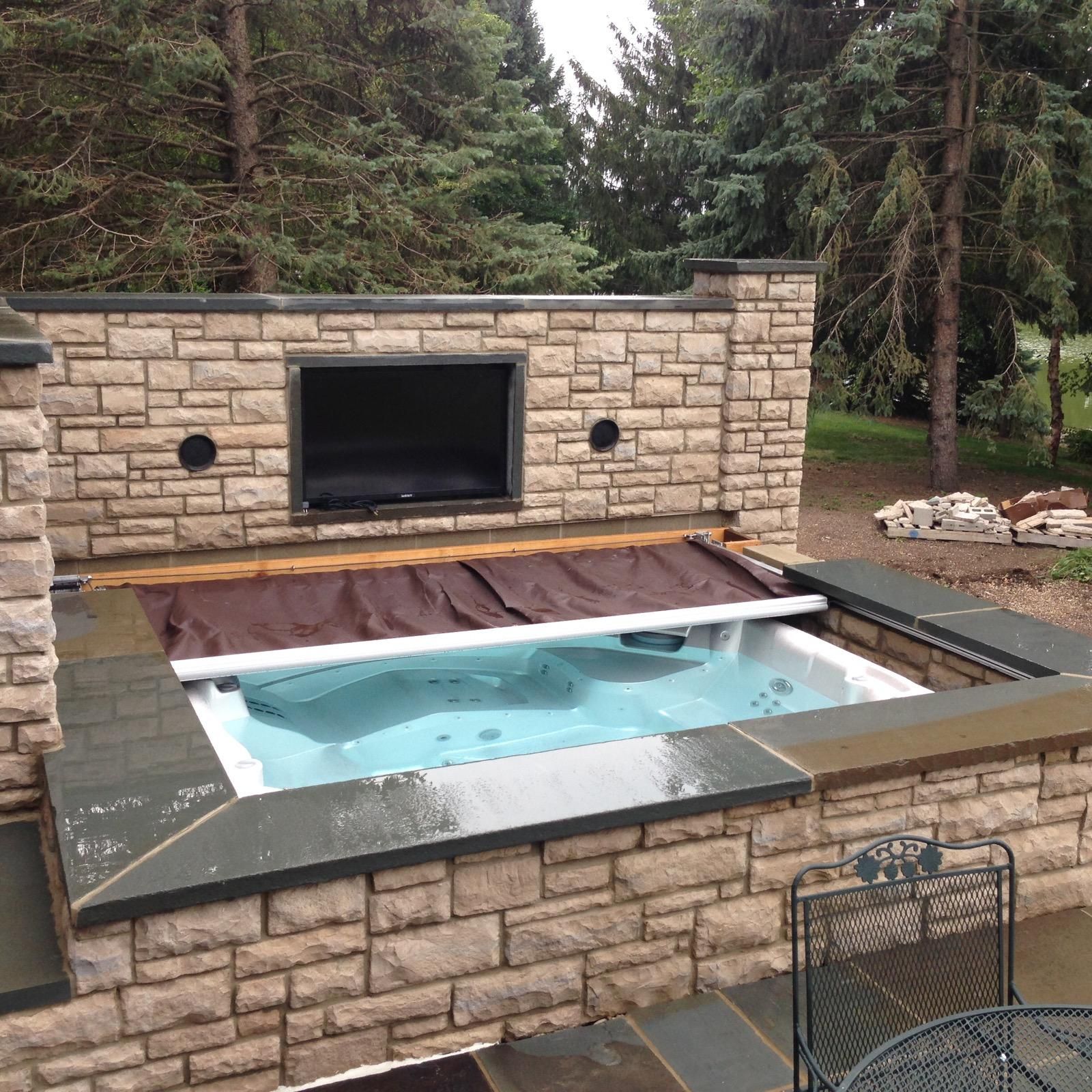 A hot tub with an automatic cover is installed next to an outdoor entertainment center in a woodsy backyard. 