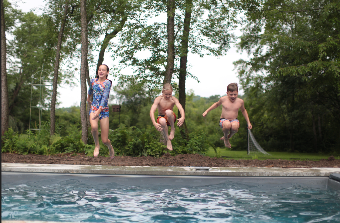 Three kids jump into a pool with a forest in the background. 