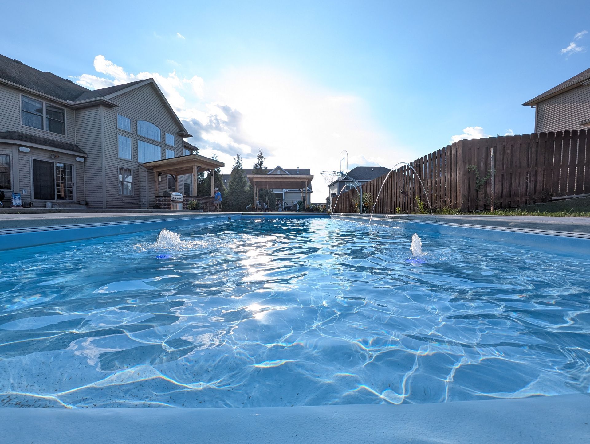 A pool is photographed at eye level with homes and fencing in the background on a beautiful, sunny day. The water is bubbling
