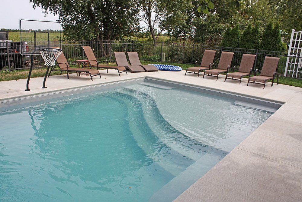 Fiberglass pool installation that has visible steps and chairs and there is a basketball net on the ledge.