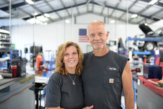 Two smiling people standing in an auto repair shop with car lifts and tools in the background