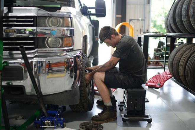 Mechanic sitting beside a silver truck, inspecting the wheel in a garage with tires stacked nearby