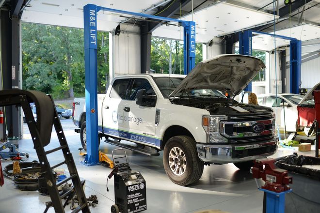 White pickup truck in an auto repair shop, hood open on a lift with tools nearby