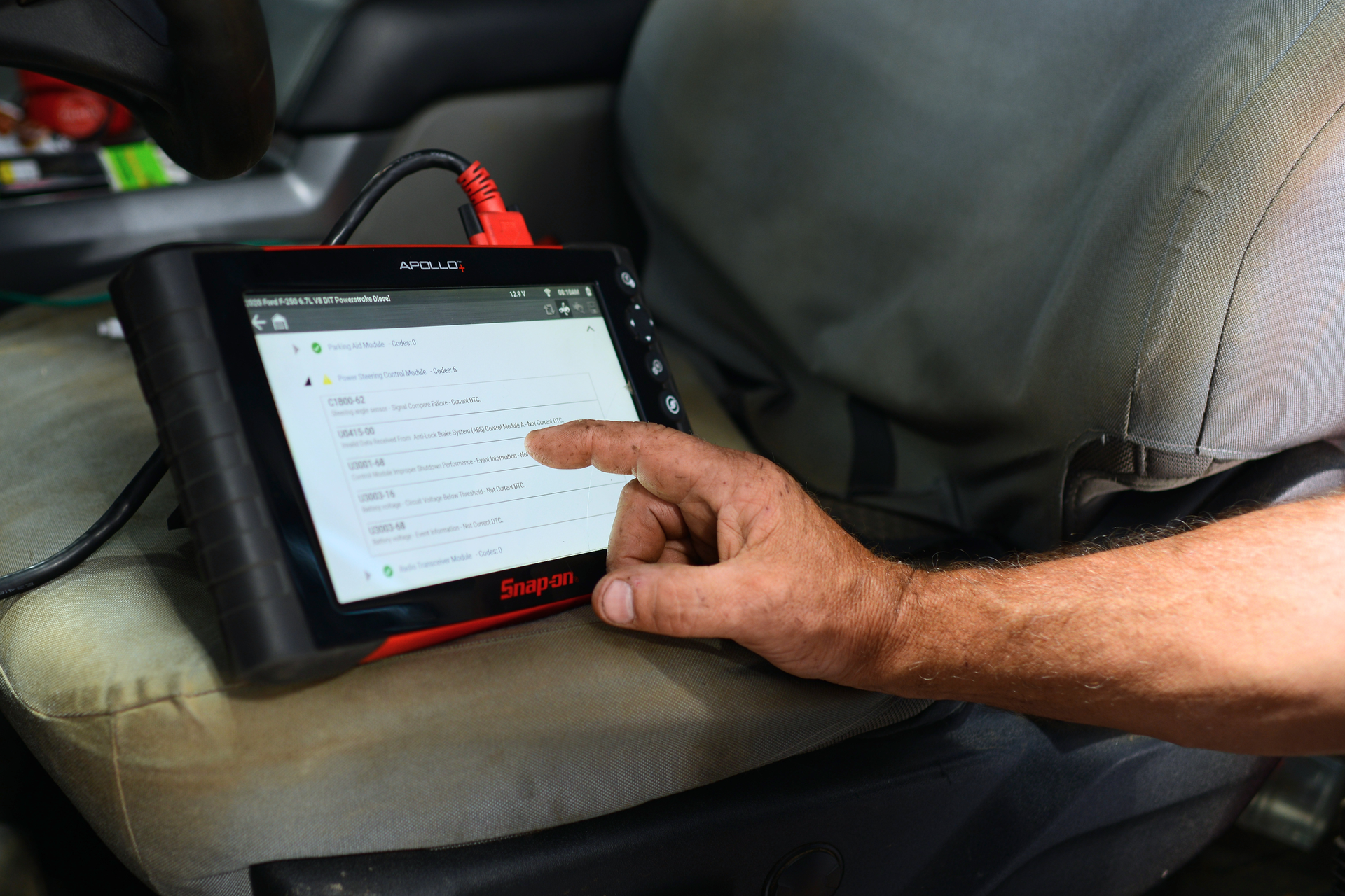 Hand touching a touchscreen tablet on a car seat, showing a menu interface