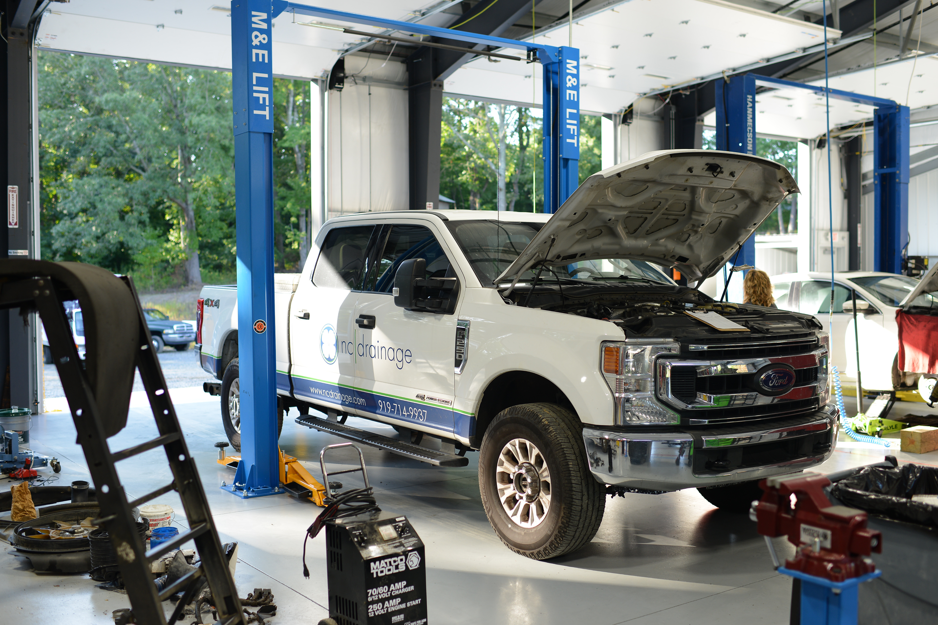 White pickup truck in an auto repair shop with hood open on a lift, surrounded by tools and equipment