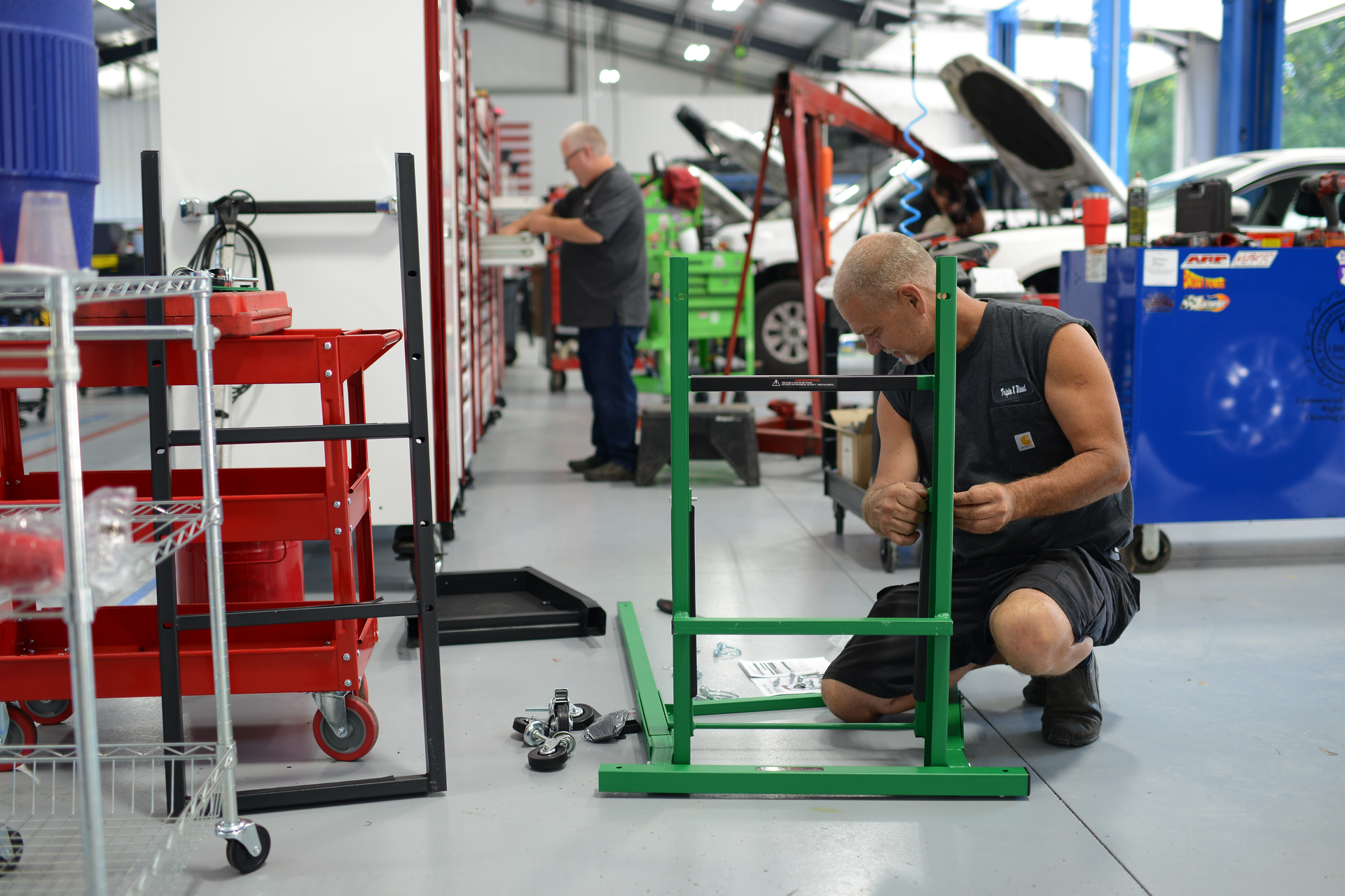 Mechanic assembling a green metal frame in an auto repair shop, with tools and a red cart nearby