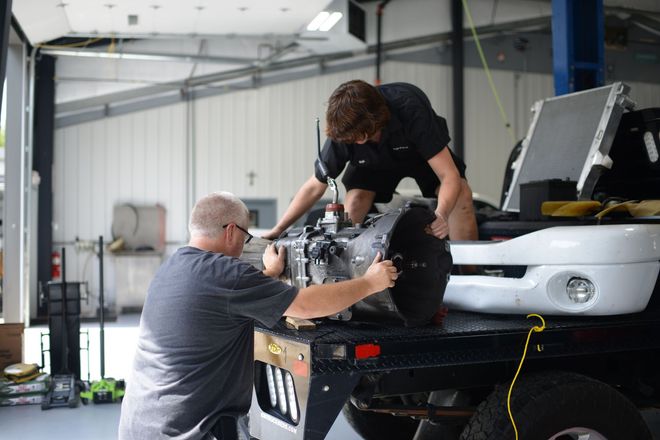 Two people installing a car bumper in a garage, with tools and equipment nearby.