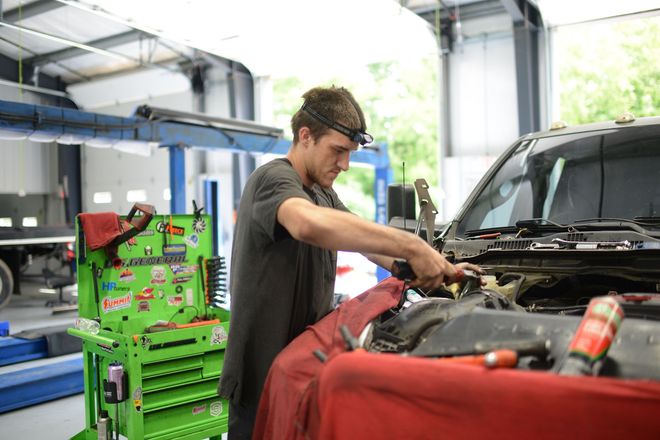 Mechanic working under an open car hood in a garage, with tools and equipment nearby.