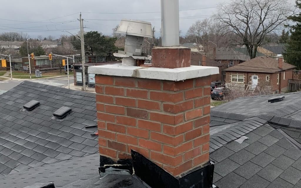 Brick chimney on a dark roof with a metal vent cap, viewed from above, with buildings in the background.