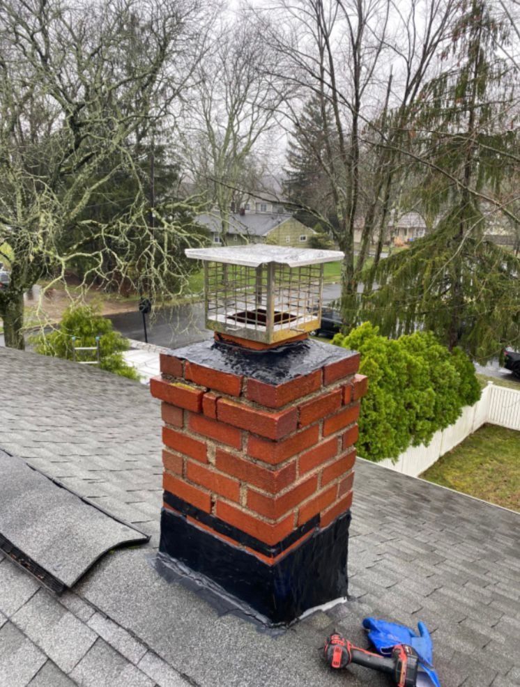 Brick chimney with a metal cap on a shingled roof, trees in background.