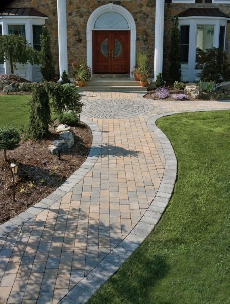 Paver walkway curves to the front door of a house with stone facade, flanked by green lawns and landscaping.