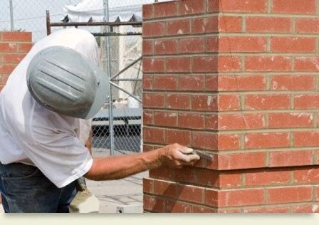 Man in hard hat using a trowel to apply mortar between red bricks on a building project.