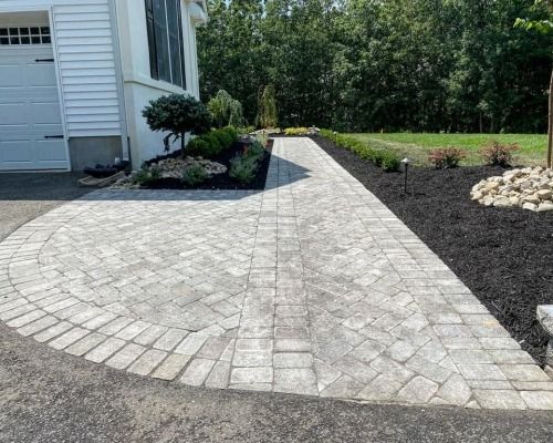 Brick pathway leading to a white house, surrounded by landscaped flower beds with dark mulch.