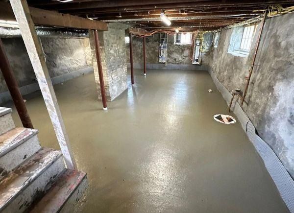 Basement with a new, smooth concrete floor. Support beams, stairs, and exposed stone walls are also visible.