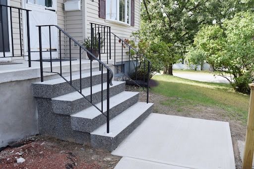 Concrete steps with black railing leading to a house entrance. Green lawn and trees in the background.