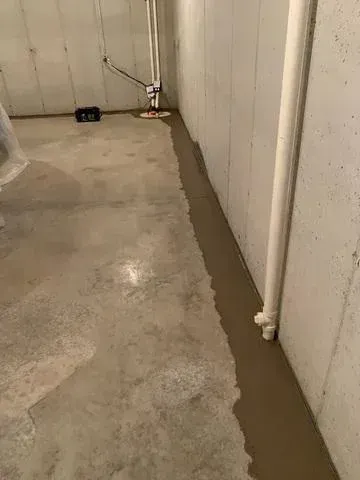Concrete basement floor with a freshly applied gray concrete strip along the wall, containing pipes and a sump pump.