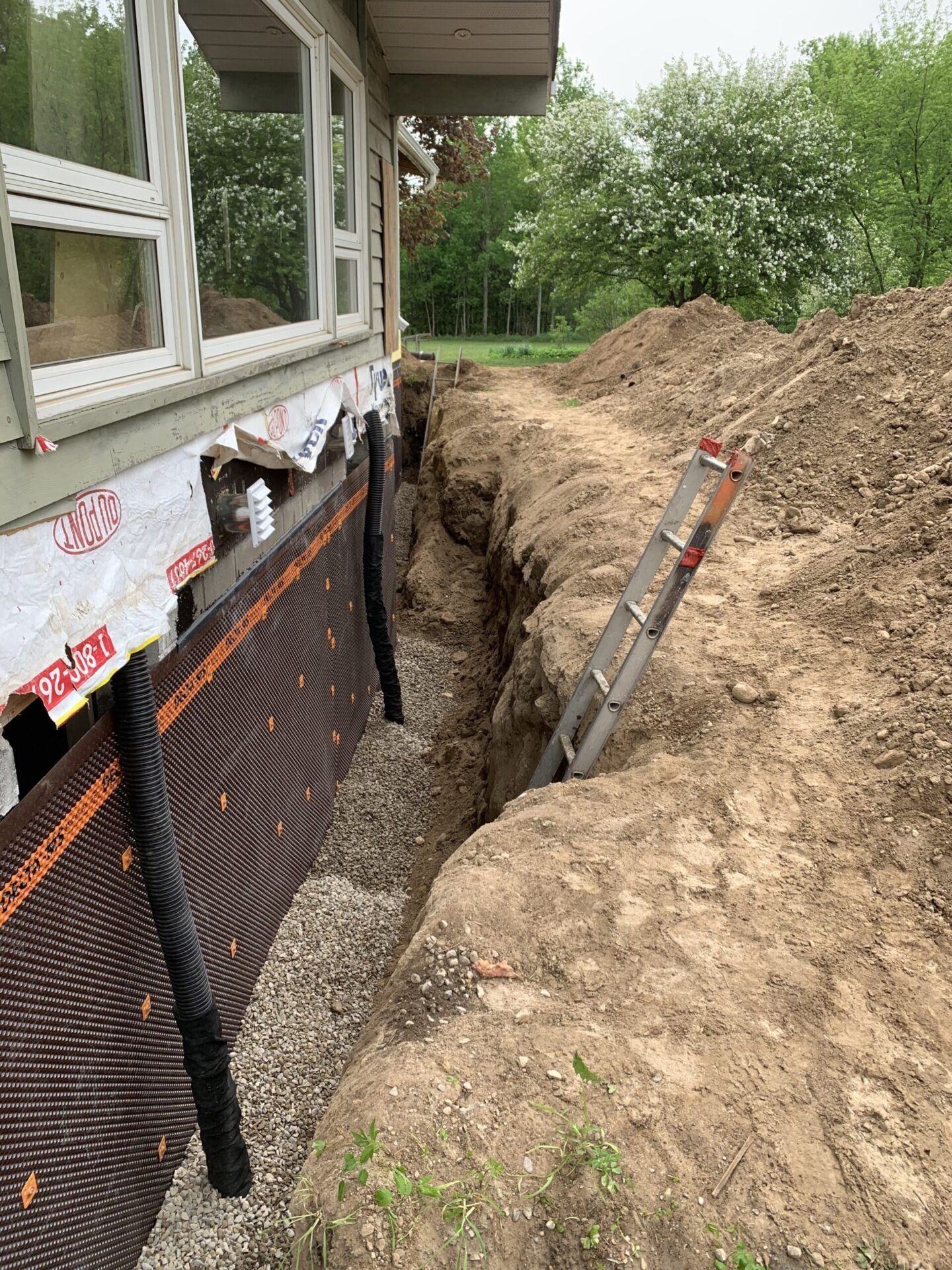 Trench dug next to a house foundation, with drainage material installed. A ladder leans against the soil.