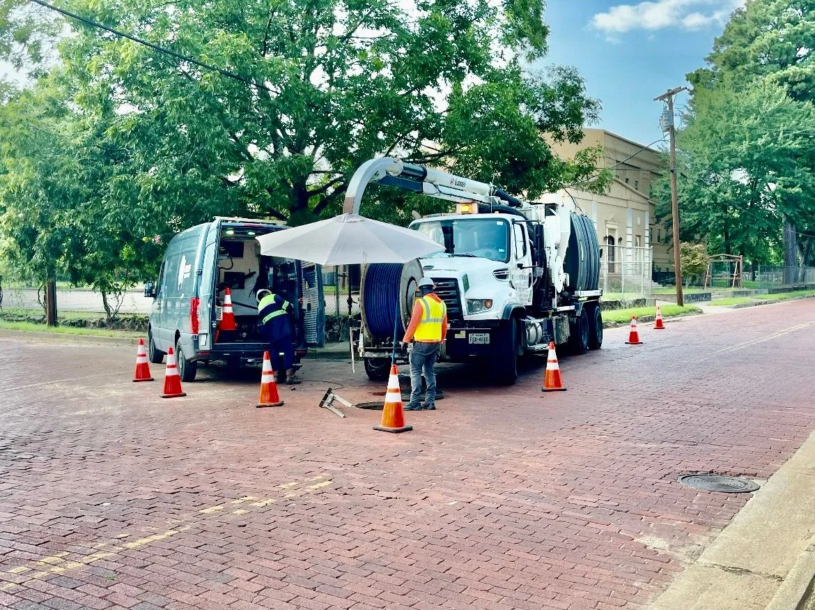 Trucks and workers on red brick road, surrounded by traffic cones. White truck with extended arm, van, and people working.