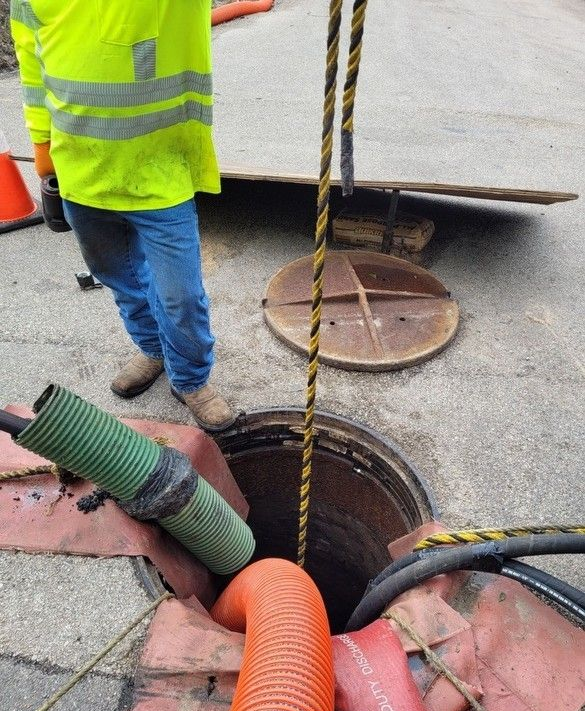 Road workers in orange vests and hard hats repairing pavement near a truck with a hose.