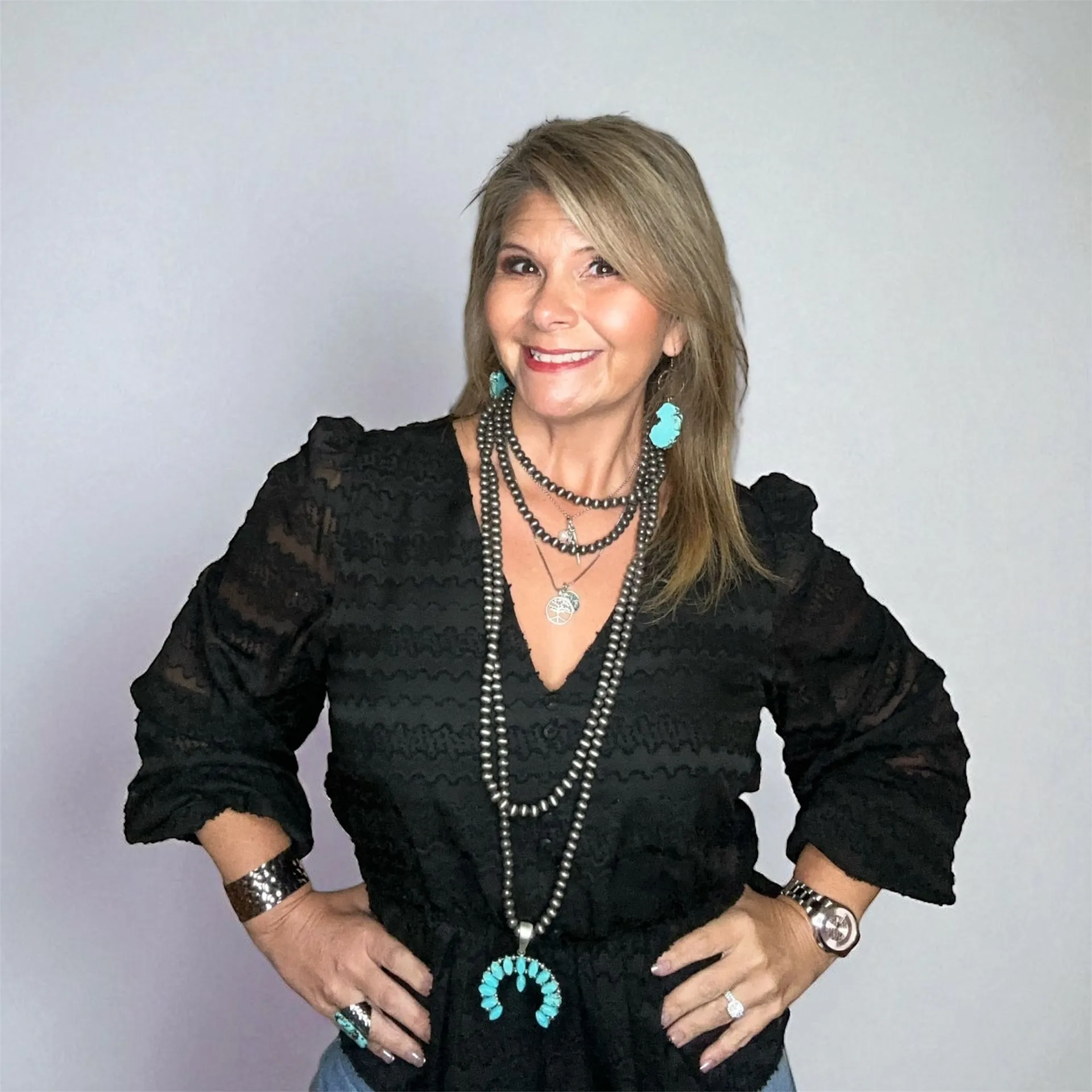Woman in black top and turquoise jewelry, hands on hips, smiling.