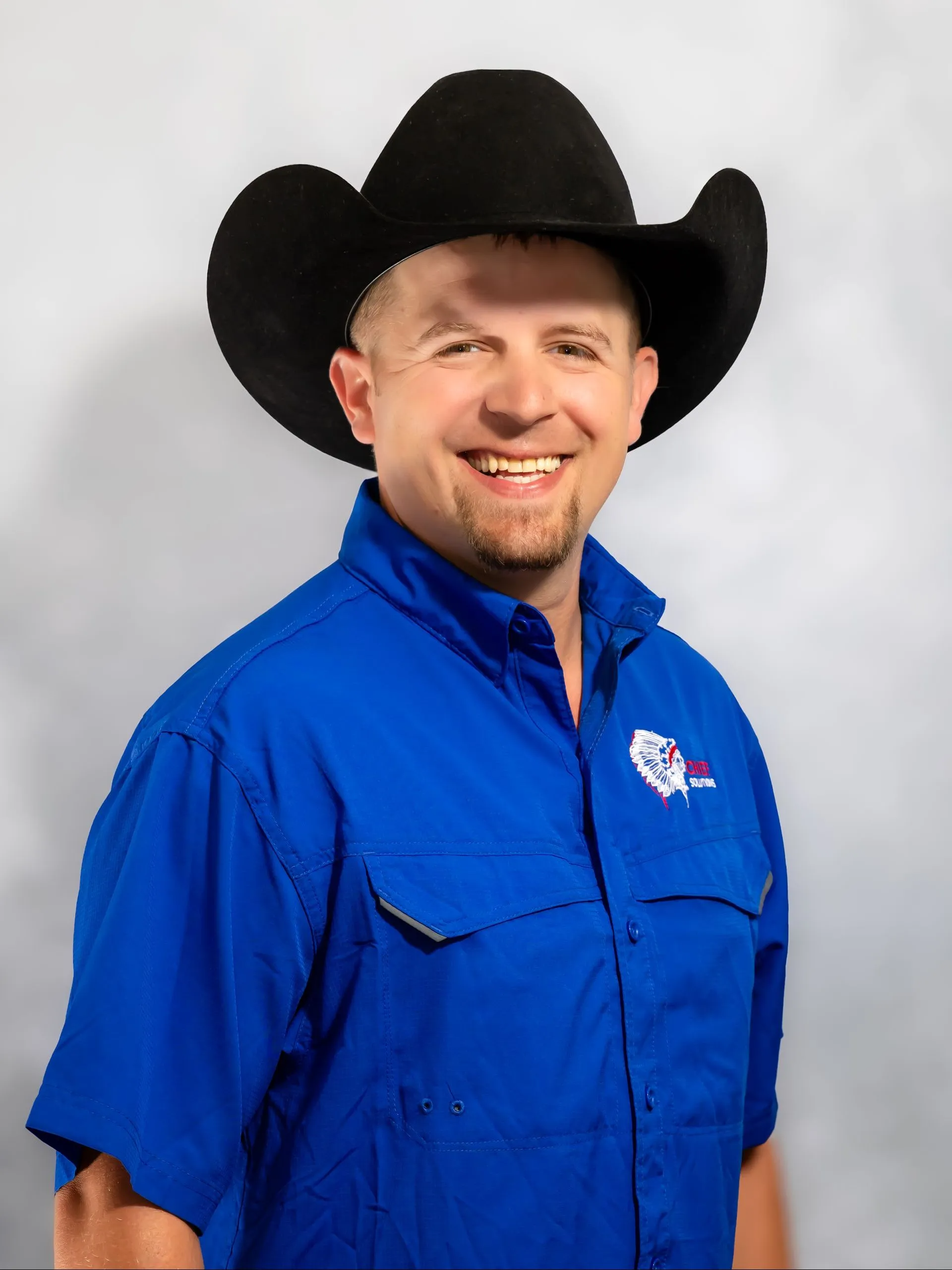 Man wearing a black cowboy hat and blue shirt smiles, with a logo on the left breast, against a light gray backdrop.