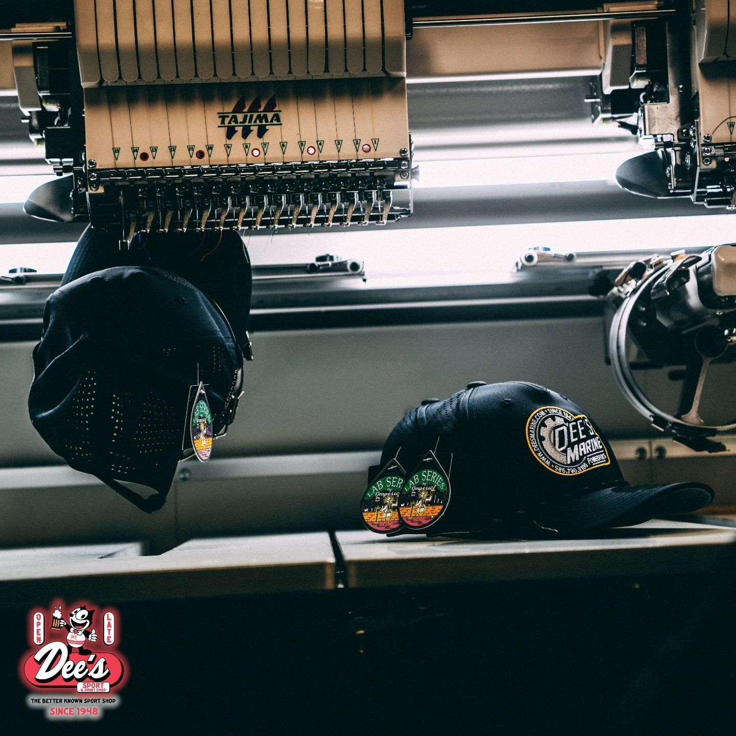 Black baseball caps under embroidery machines in a shop, with a red “Dee’s” logo in the corner