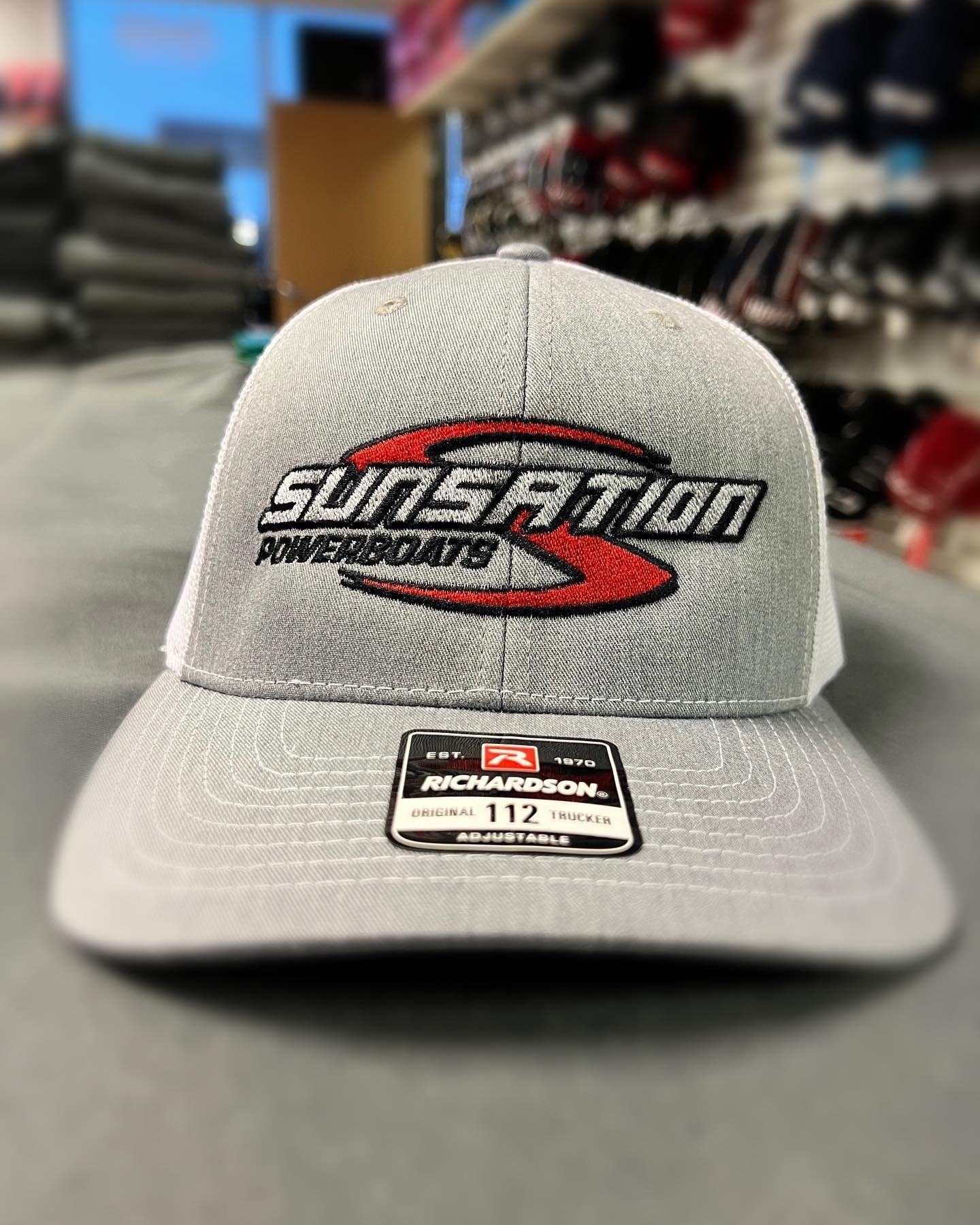 Gray SunSation baseball cap with a black-and-red logo and a McArthur patch, displayed in a store.