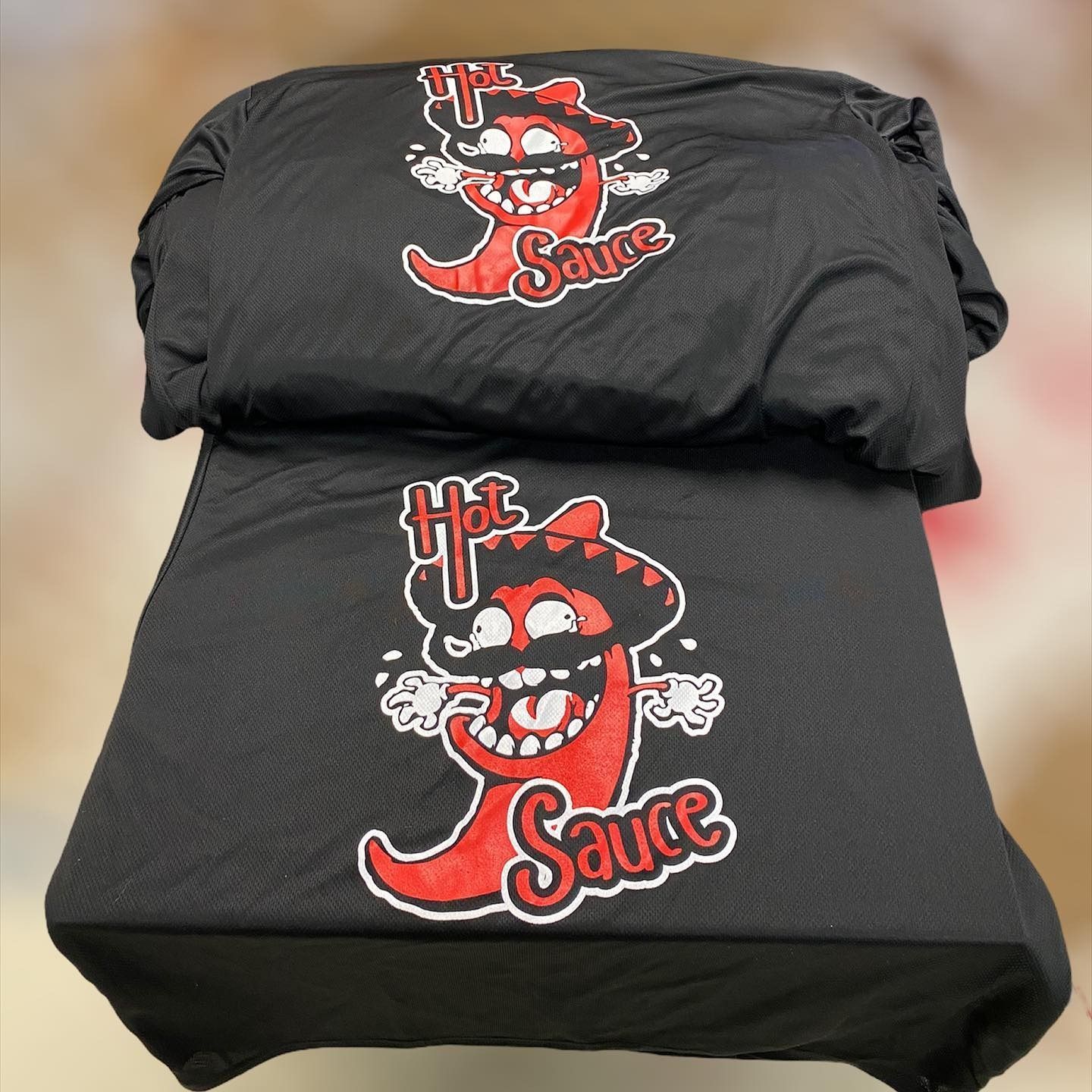 Two black folded shirts with a red-and-white hot sauce cartoon logo on the front