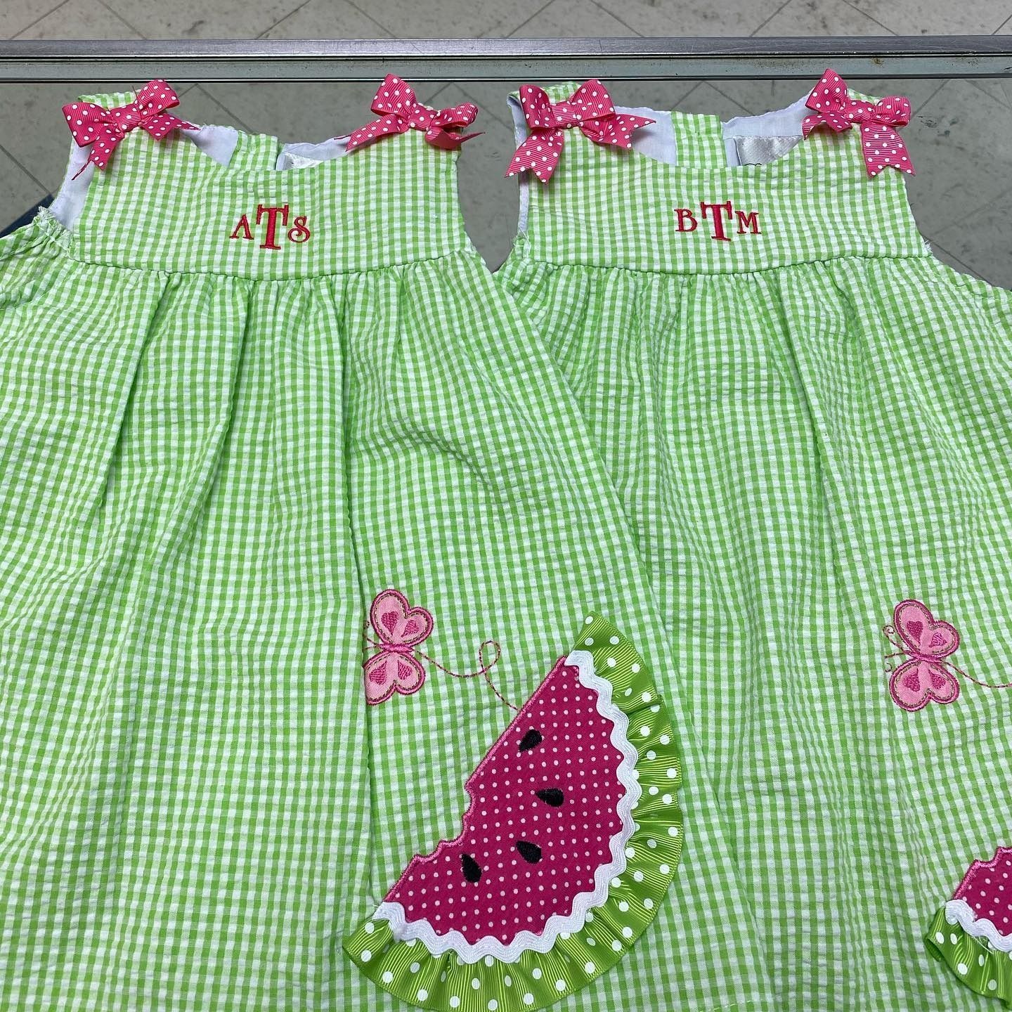 Two green gingham children’s dresses with pink bows and watermelon appliqués on a white background