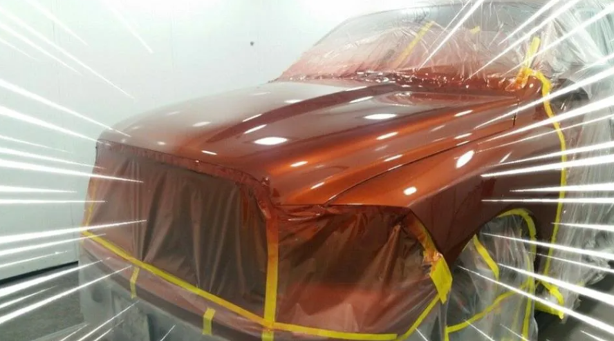 Orange car masked with plastic and tape, being spray painted in a workshop