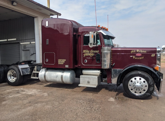 Maroon semi-truck parked beside a warehouse with silver fuel tanks and chrome wheels