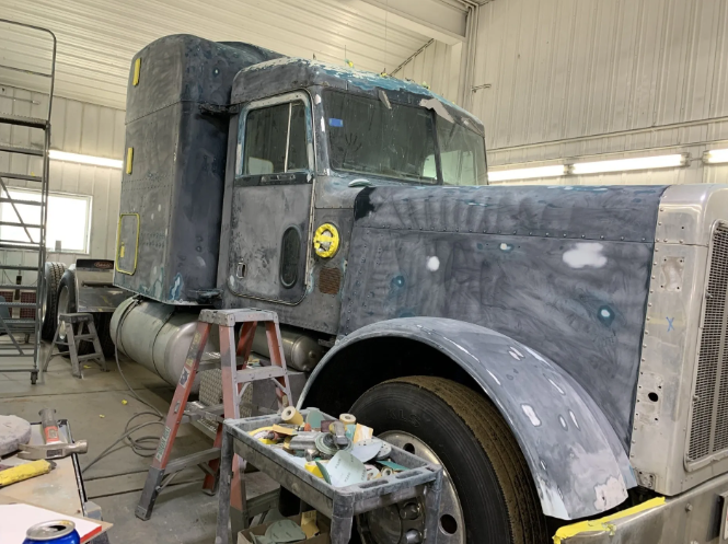 Old black semi-truck in a garage, partially sanded and covered for restoration work.