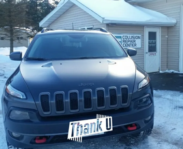 Black Jeep parked in front of a collision repair shop, with a novelty “thank U” front plate.
