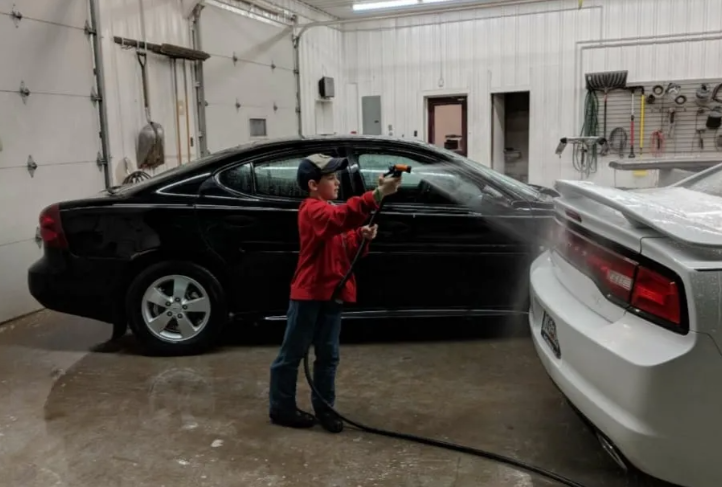 Person washing a black car beside a white car in a garage.